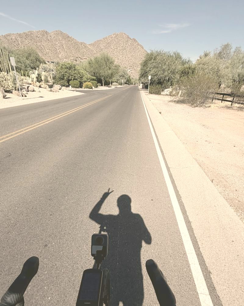 A rider&rsquo;s shadow on the road, one hand up off the bars, mountains ahead.