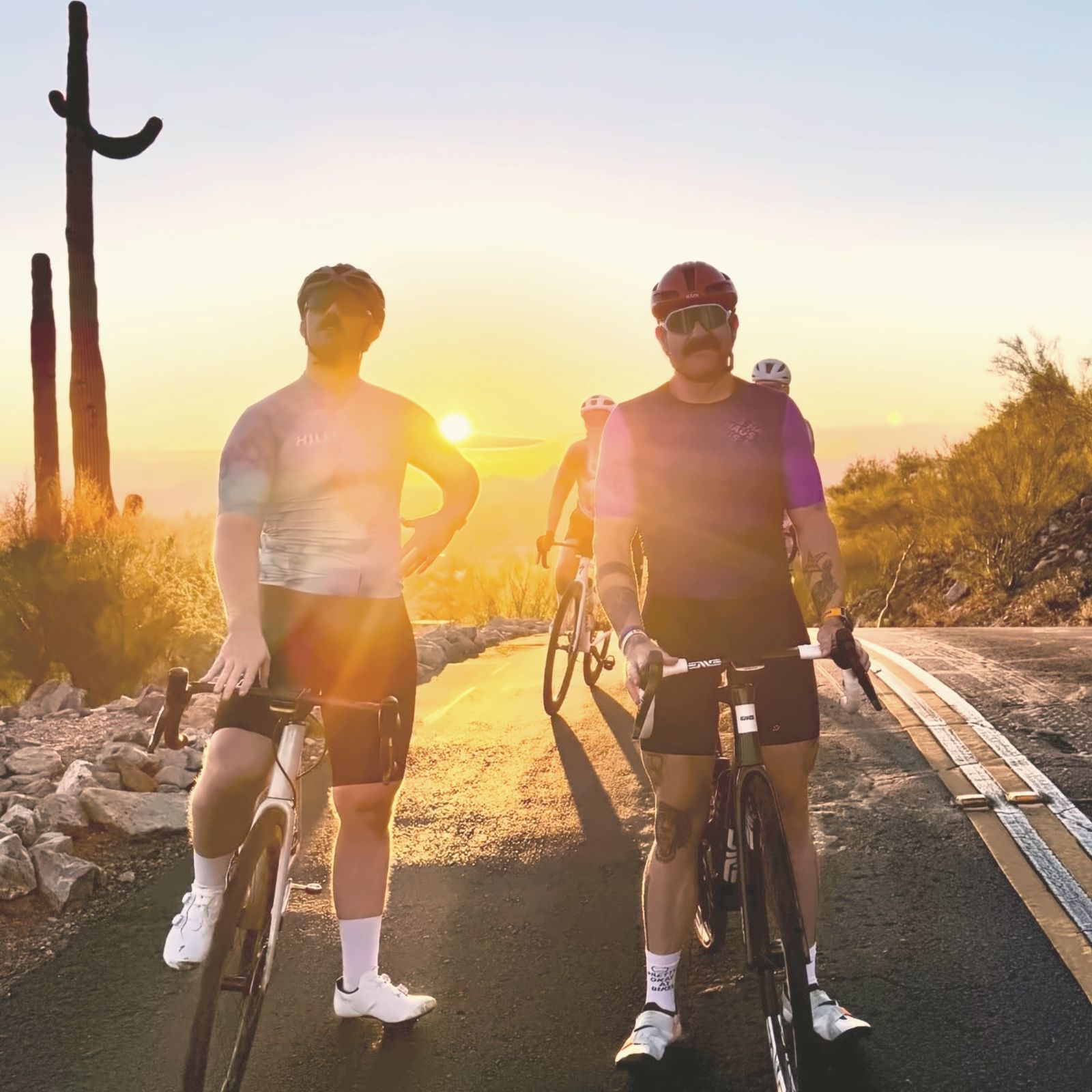 Two riders stopped on a desert road at sunset, saguaros behind them, the sun sitting on the horizon.