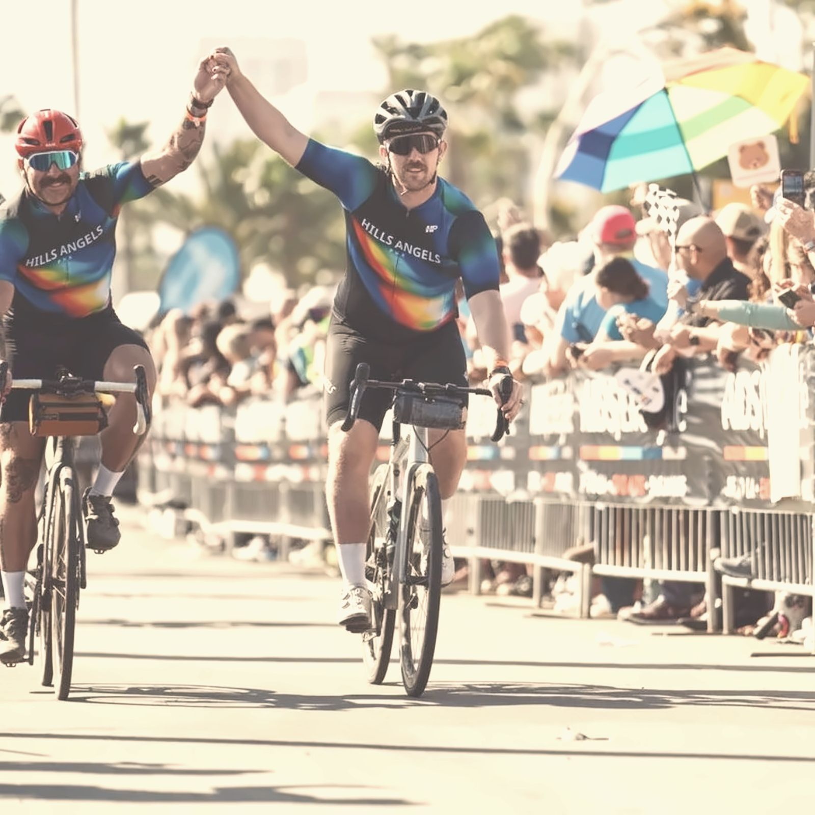 Two riders crossing a finish line with their hands joined in the air, a rainbow umbrella in the crowd.